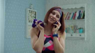 Spor kıyafetleri içinde, elinde dambıllar ve cep telefonu olan 30 'lu yaşlardaki genç bir kadın hücresinde ağır çekimde konuşuyor.. 