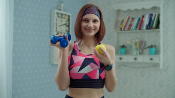 Young 30s woman in sportswear with dumbbells and apple fruit in hands smiling looking at camera at home in slow motion. Fitness, yoga and sport at home concept. — Stock Video