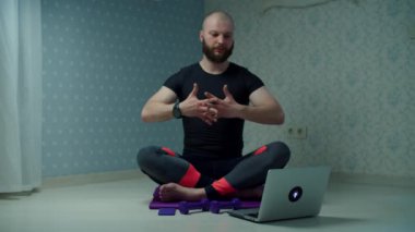 Spor giyimli 30 'lu yaşlarda yoga yapan ve spor paspası üzerinde egzersiz yapan sakallı bir adam evde dizüstü bilgisayar kullanarak spor yapıyor. Adam yavaş çekimde halterlerle spor yapıyor.. 