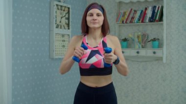 Spor kıyafetli 30 'lu yaşlardaki genç bir kadın evde spor egzersizleri yapıyor. Kadın evde el egzersizi yapıyor. Ağır çekimde kameraya bakıyor.. 
