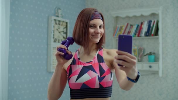 Młoda kobieta po trzydziestce w odzieży sportowej z hantlami i telefonem komórkowym w rękach, robiąca zdjęcie selfie i uśmiechająca się do domu w zwolnionym tempie. — Wideo stockowe