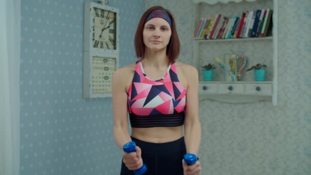 Młoda kobieta po trzydziestce w odzieży sportowej z hantlami ćwicząca fitness w domu. Kobieta robi ćwiczenia rąk w domu patrząc na kamerę w zwolnionym tempie. — Wideo stockowe