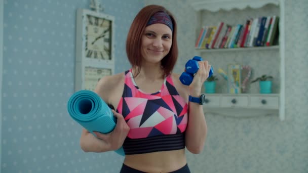 Νεαρή 30άρα γυναίκα με αθλητικά ρούχα με αλτήρες και στρώμα γυμναστικής στα χέρια χαμογελώντας κοιτάζοντας την κάμερα στο σπίτι σε αργή κίνηση. Άσκηση, γιόγκα και αθλητισμός στο σπίτι έννοια. — Αρχείο Βίντεο