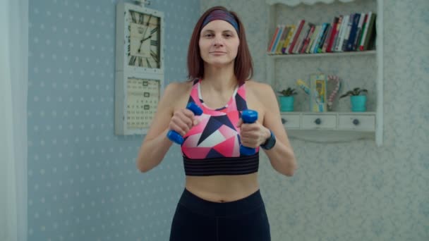 Młoda kobieta po trzydziestce w odzieży sportowej z hantlami ćwicząca fitness w domu. Kobieta robi ćwiczenia rąk w domu patrząc na kamerę w zwolnionym tempie. — Wideo stockowe