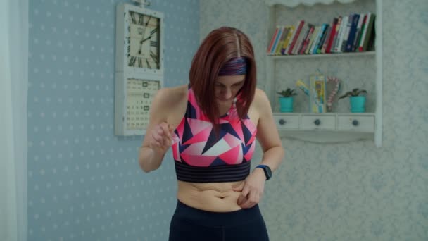 Νεαρή 30άρα γυναίκα σε αθλητικά ρούχα δυστυχισμένη με χοντρή κοιλιά και θέλει να χάσει βάρος στο σπίτι σε αργή κίνηση. Γυμναστική, γιόγκα και χαλαρό βάρος στο σπίτι έννοια. — Αρχείο Βίντεο