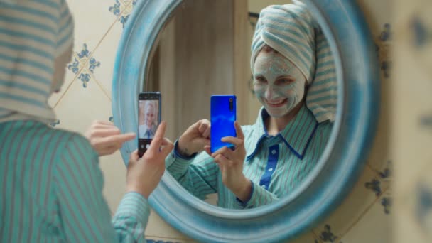 Mujer joven de 30 años con toalla en la cabeza y máscara de cosméticos azules en la cara hablando por videollamada en el baño en casa. Reflejo espejo de mujer en máscara azul con teléfono móvil en las manos — Vídeos de Stock