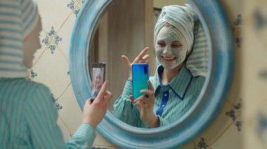 Kafasında havlu ve yüzünde mavi kozmetik maske olan 30 'lu yaşlardaki genç bir kadın evdeki banyoda video konuşmalarıyla konuşuyor. Elinde cep telefonu olan mavi maskeli bir kadının ayna yansıması. 