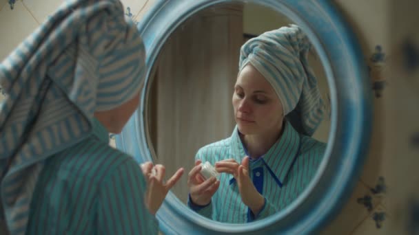 Mujer joven de 30 años con toalla en la cabeza aplicando crema hidratante para la cara en el baño azul en casa. Reflejo del espejo de la mujer haciendo procedimientos de belleza en casa. De cerca. — Vídeos de Stock