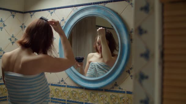 Dertiger jaren vrouw in handdoek verven haar haar thuis met borstel en kleurstof in spiegelreflectie. Brunette vrouw met naakte schouders in badkamer thuis het maken van spa. — Stockvideo