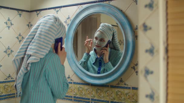 Jeune femme des années 30 avec serviette sur la tête et masque cosmétique bleu sur le visage parlant par téléphone portable dans la salle de bain à la maison. Reflet miroir de la femme en masque bleu avec téléphone portable dans les mains — Video