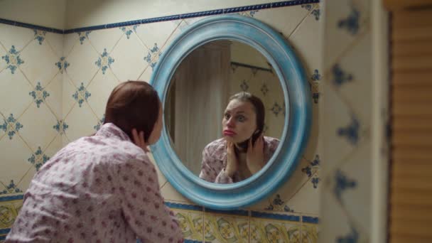 Jonge dertiger jaren vrouw wassen gezicht gevoel hoofdpijn in de badkamer in de voorkant van spiegel met slechte make-up en verwend haar styling. Alcohol bedwelmde vrouw in roze shirt in blauwe ronde spiegel reflectie. — Stockvideo