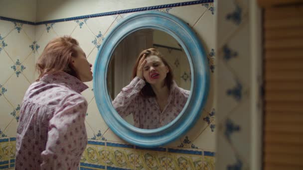 Молодая 30-летняя женщина чувствует головную боль в ванной комнате перед зеркалом с плохим макияжем и испорченной прической. Опьяненная алкоголем женщина в розовой рубашке в синем круглом зеркале . — стоковое видео