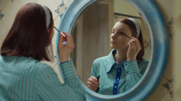 Young 30s woman in blue shirt applying mascara on eyes in blue bathroom at home. Mirror reflection of lady doing make up at home. — Stock Video