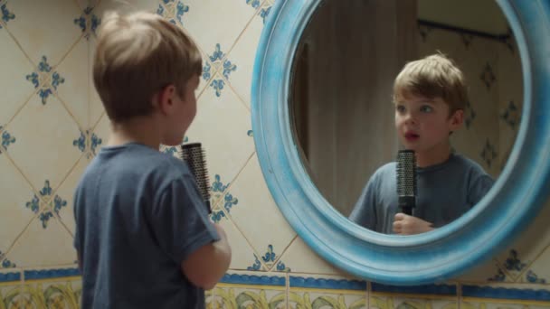 Niño rubio preescolar cantando en cepillo de pelo y bailando en espejo reflejo en casa en el baño. Niño feliz divirtiéndose en casa . — Vídeo de stock