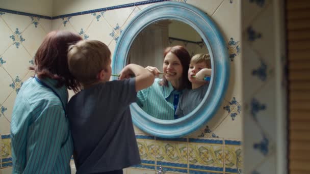 Joven madre de 30 años con hijo preescolar cepillándose los dientes en el baño en casa. Familia feliz en espejo reflexión haciendo rutina de la mañana juntos . — Vídeos de Stock