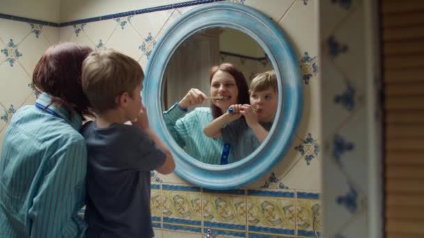 Jeune mère des années 30 avec un fils d'âge préscolaire se brossant les dents dans la salle de bain à la maison. Famille heureuse dans le reflet miroir faire la routine du matin ensemble . — Video