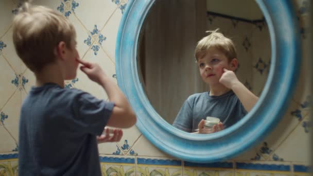 Blond preschooler boy applying moisturizing cream on his face in mirror reflection at home in bathroom. Happy kid having fun at home. — Stock Video