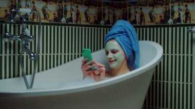30 'lu yaşlarda esmer bir kadın cep telefonuyla sohbet ediyor evdeki yeşil banyoda köpüklü küvette oturuyor. Yüzünde kozmetik maske olan bir kadın küvette cep telefonuyla rahatlıyor.. 