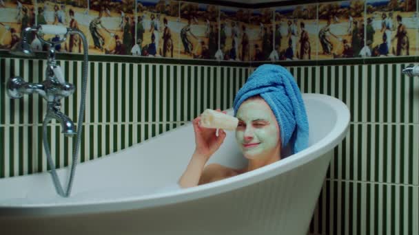 Jeune femme des années 30 dans la baignoire avec mousse tenant éponge écologique dans les mains et souriant en regardant la caméra à la salle de bain verte à la maison. Femme avec masque cosmétique vert sur le visage faisant des procédures de bain . — Video