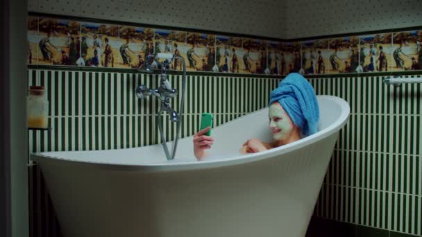 Brunette 30s woman taking picture using mobile phone sitting in bathtub with foam at green bathroom at home. Woman with cosmetic mask on face relaxing with cell phone in bathtub. — Stock Video