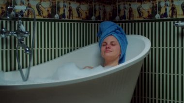 30 'lu yaşlardaki genç bir kadın evdeki yeşil banyoda köpükle banyo yapıyor. Kafasında mavi havlu olan bir kadın küvette dinleniyor. Kapat.