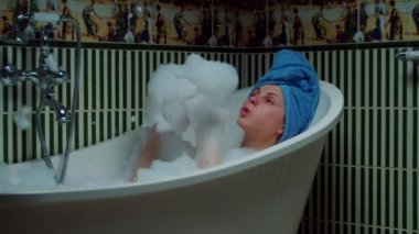 30 'lu yaşlardaki genç bir kadın evdeki yeşil banyoda banyo yapıyor ve köpükle oynuyor. Kafasında mavi havlu olan kadın küvette dinleniyor.. 