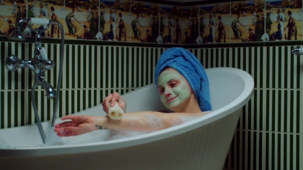 Mujer joven de 30 años lavándose las manos con esponja ecológica en la bañera con espuma en el baño verde en casa. Mujer con máscara cosmética verde en la cara haciendo procedimientos de baño. De cerca. — Vídeos de Stock