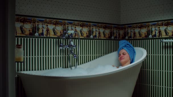 Young 30s woman taking bath with foam at green bathroom at home. Woman with blue towel on head relaxing in bathtub. — Stock Video