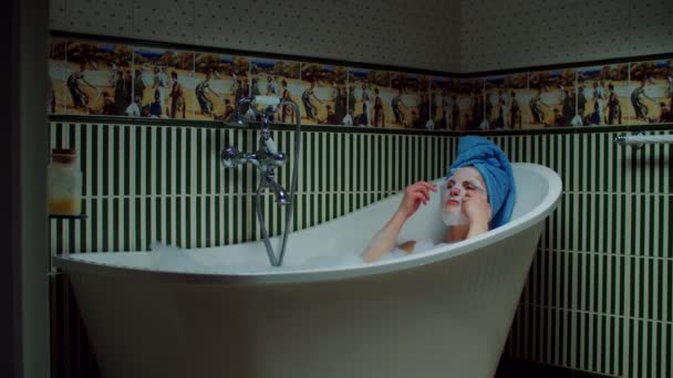 Młoda kobieta po trzydziestce nakładająca kosmetyczną maseczkę na twarz, siedząca w wannie z pianką w zielonej łazience w domu. Kobieta z niebieskim ręcznikiem na głowie relaksująca się w wannie. Home koncepcja spa. — Wideo stockowe