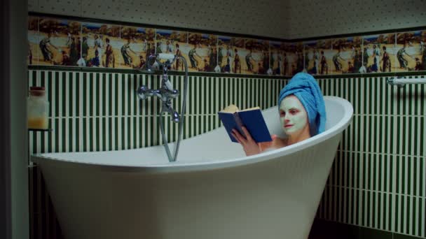 Mulher morena dos anos 30 lendo livro de papel na banheira com espuma no banheiro verde em casa. Mulher com máscara cosmética no rosto gosta de ler livro na banheira . — Vídeo de Stock
