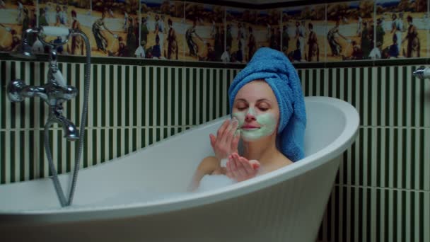 Junge Frau in den 30er Jahren, die kosmetische Gesichtsmaske aufträgt, sitzt in der Badewanne mit Schaum im grünen Badezimmer zu Hause. Frau mit blauem Handtuch auf dem Kopf entspannt in Badewanne. Home-Spa-Konzept. Nahaufnahme — Stockvideo