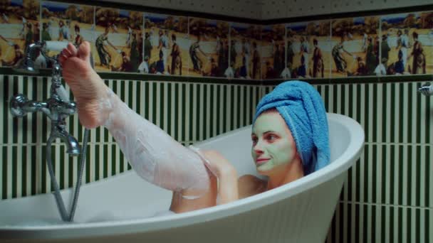 Jeune femme des années 30 se rasant les jambes dans la baignoire avec de la mousse à la salle de bain verte à la maison. Femme avec masque cosmétique vert sur le visage faisant des procédures de bain. Gros plan — Video