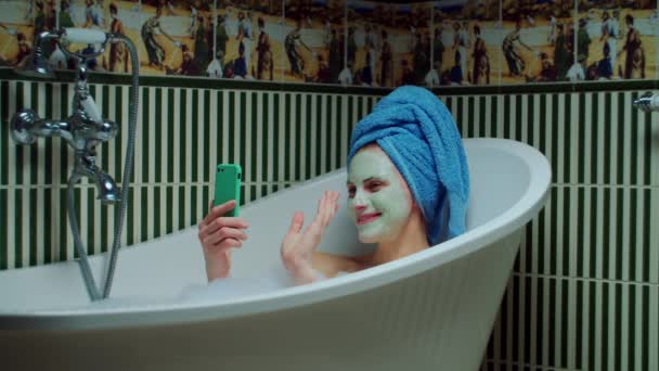 Morena mujer de 30 años tomando fotos utilizando el teléfono móvil sentado en la bañera con espuma en el baño verde en casa. Mujer con máscara cosmética en la cara relajante con teléfono celular en la bañera . — Vídeo de stock