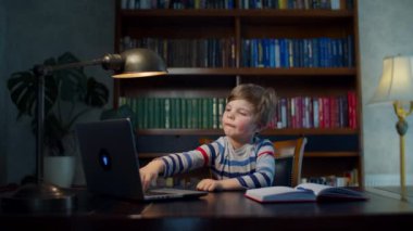 Anaokulu öğrencisi laptopla online okul ödevi yapıyor. Çocuk evdeki ahşap masada oturuyor ve internetten ders çalışıyor. Egzersiz kitabına kalemle yazıyor.. 
