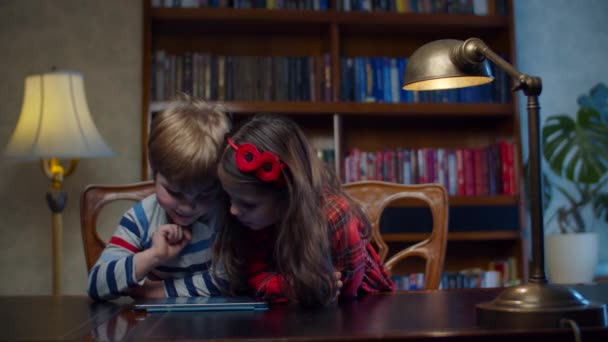 Двое дошкольников играют в игры на планшетном компьютере дома. Братья и сестры смеются вместе, используя приложение на гаджете. Счастливые мальчик и девочка играют онлайн . — стоковое видео