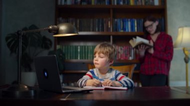 Anaokulu öğrencisi dizüstü bilgisayarla ödev yapıyor, annesi de arkasında kağıt okuyor. Çocuk evde online çalışıyor, kalemle egzersiz defterine yazıyor. Ayakta duran ve kitap okuyan kadın