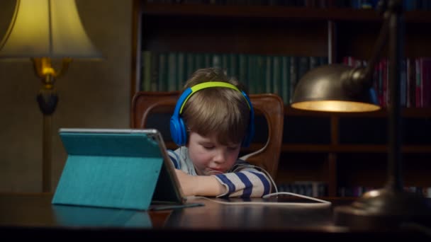 Menino pré-escolar em fones de ouvido coloridos cansado do computador tablet. Criança empurrando para trás gadget sentado na mesa com lâmpada. Criança perturbada cansada de tablet digital . — Vídeo de Stock