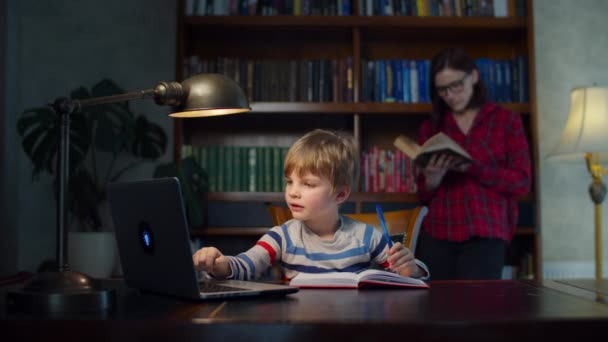 Kleuterjongen die online schoolhuiswerk maakt met laptop, moeder die het papierboek leest. Hij studeert thuis online, schrijft op met pen in het oefenboek. Vrouw staande en leesboek — Stockvideo