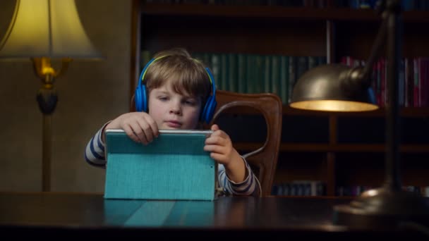 Niño preescolar en auriculares de color usando tableta en casa. Niño viendo contenido en línea en gadget sentado en el escritorio con la lámpara en cámara lenta . — Vídeos de Stock