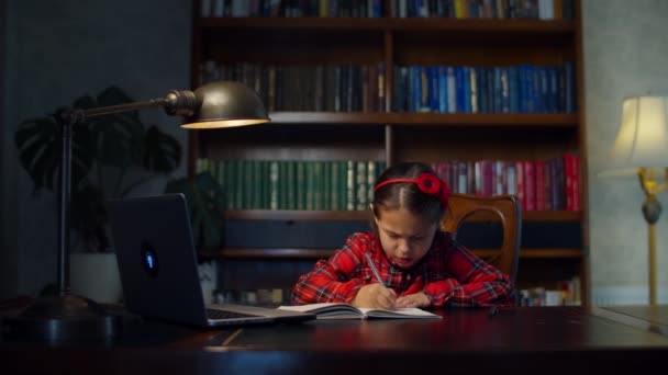 Chica de la escuela en vestido rojo haciendo tarea de la escuela en línea con el ordenador portátil en casa. Niño sentado en el escritorio de madera y estudiando en línea, escribiendo con pluma en el libro de ejercicios. Proceso de educación en línea . — Vídeo de stock