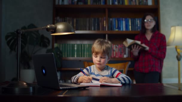 ノートパソコンでオンライン学校の宿題を作る未就学児の少年、背後にある母親の読書紙の本。子供は家でオンラインで勉強し、練習本のペンで書き留める。立って本を読む女 — ストック動画
