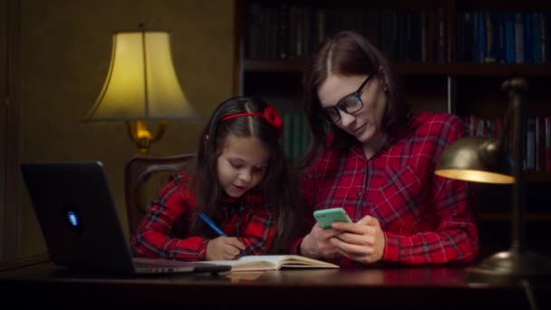 School meisje maakt huiswerk met jonge moeder uit de 30 met behulp van mobiele telefoon thuis. Een jongen die naar een laptop kijkt en opschrijft in een notitieboekje. Online onderwijsproces. Familie kijken mam en dochter. — Stockvideo