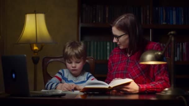 Vorschulkind macht Online-Hausaufgaben mit einer jungen Mutter aus den 30er Jahren zu Hause. Kind schaut sich Lehrbücher an und notiert in Notizbuch. Home Online-Bildungsprozess. — Stockvideo