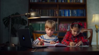 Evde online ödev yapan iki anaokulu çocuğu. Kardeşler dizüstü bilgisayara bakarak kalemlerle birlikte yazıyorlar. Çevrimiçi ev eğitimi süreci. 