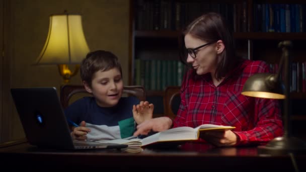 Uczeń odrabiający lekcje z młodą trzydziestolatką w domu. Dzieciak przegląda podręcznik i zapisuje w notatniku. Proces edukacji domowej. — Wideo stockowe