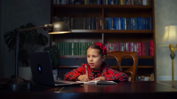 School meisje in rode jurk het maken van online school huiswerk met laptop thuis. Kind zit aan het houten bureau en studeert online, schrijft op met pen in het oefenboek. Online onderwijsproces. — Stockvideo
