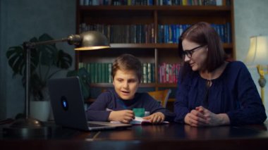Liseli çocuk evde 30 'lu yaşlardaki genç annesiyle internette ödev yapıyor. Çocuk dizüstü bilgisayara bakıyor ve not defterine yazıyor. Evde çevrimiçi eğitim süreci.