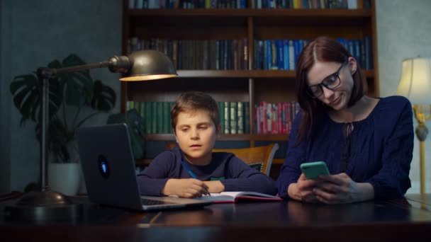 Menino da escola fazendo trabalhos de casa on-line com a jovem mãe dos anos 30 usando telefone celular em casa. O miúdo a olhar para o portátil e a escrever no caderno. Processo de educação on-line em casa . — Vídeo de Stock