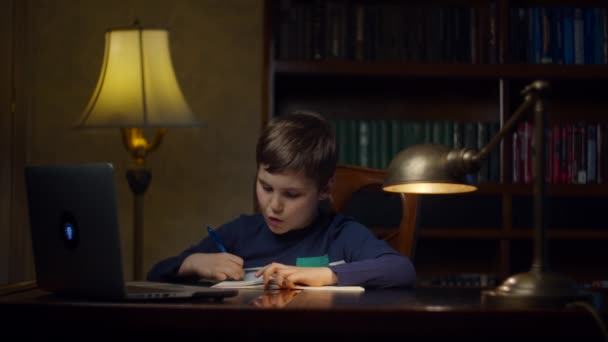 家庭でラップトップでオンライン学校の宿題を作る学校の少年。子供は木製の机に座ってオンラインで勉強し、練習本のペンで書き留める。オンライン教育プロセス. — ストック動画