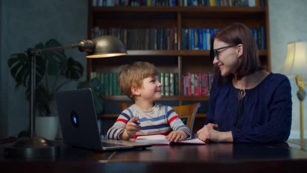 Niño preescolar haciendo tarea en línea con la madre joven de 30 años en casa. Un chico escribiendo en un libro de ejercicios. Proceso de educación en línea . — Vídeos de Stock